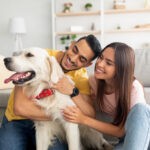 Portrait of happy multiracial couple scratching their pet dog, sitting on floor at home
